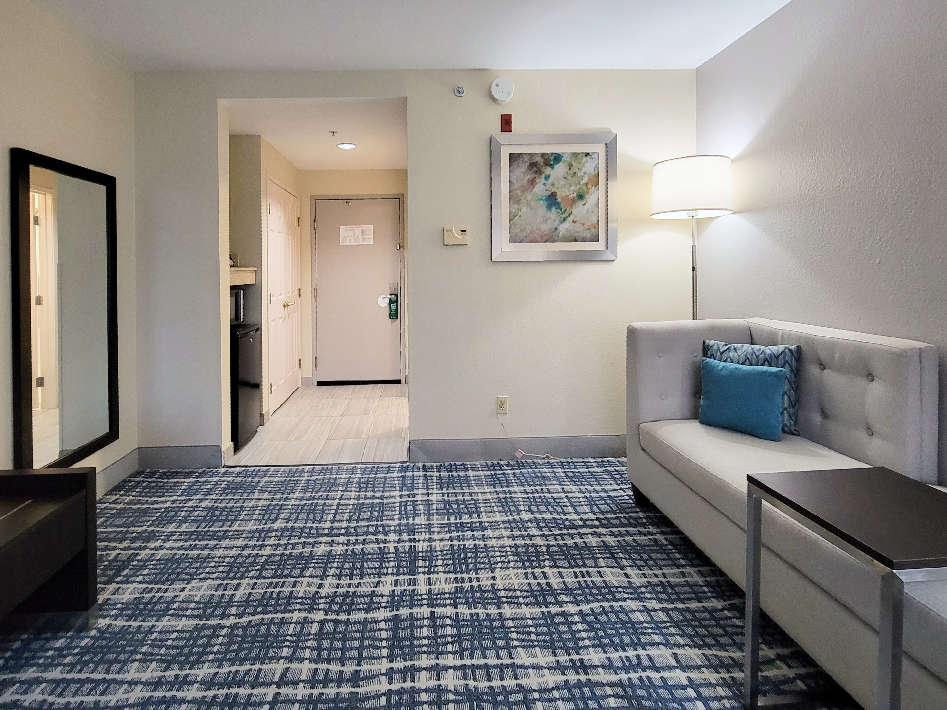 Une chambre d’hôtel avec une moquette à motifs, un canapé avec un coussin bleu, une table de chevet, une lampe et une œuvre d’art encadrée accrochée au mur.