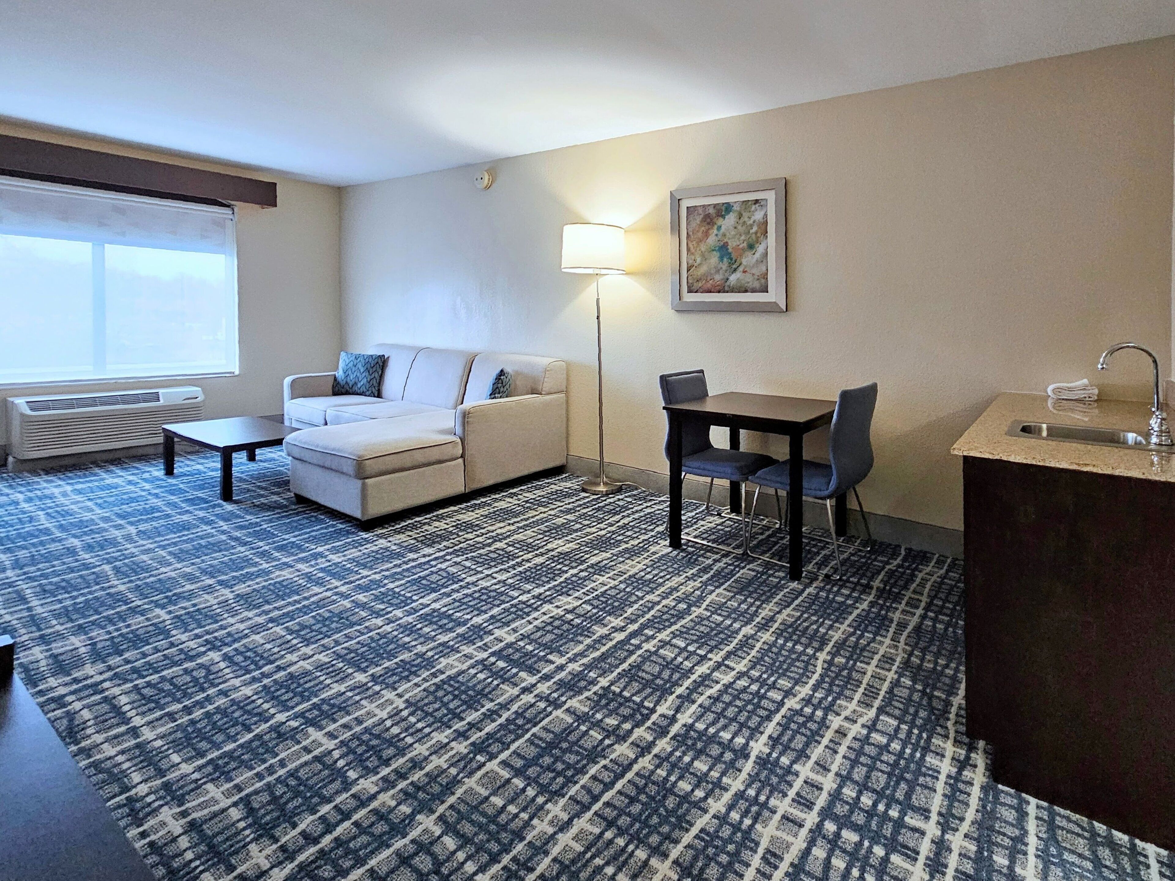 A hotel room with a sofa, a small table, a desk with chairs, a lamp, and a sink.