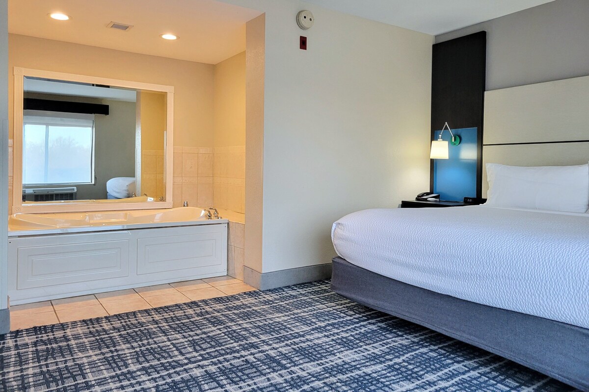 A hotel room with a large bed, a double sink, a mirror, and a bedside table with a lamp.