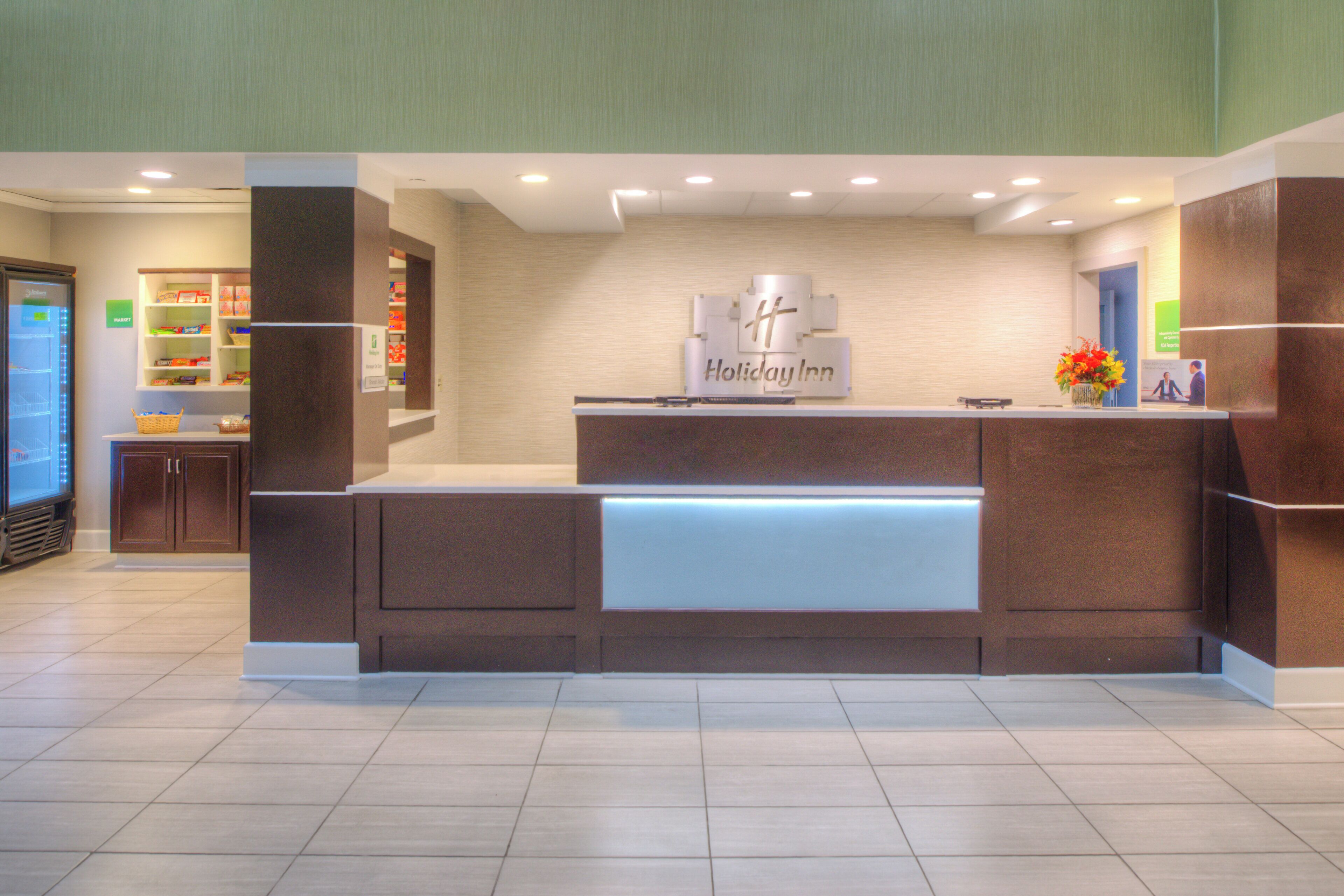 A reception desk with a Holiday Inn sign, a market area with shelves stocked with products, and a refrigerator.