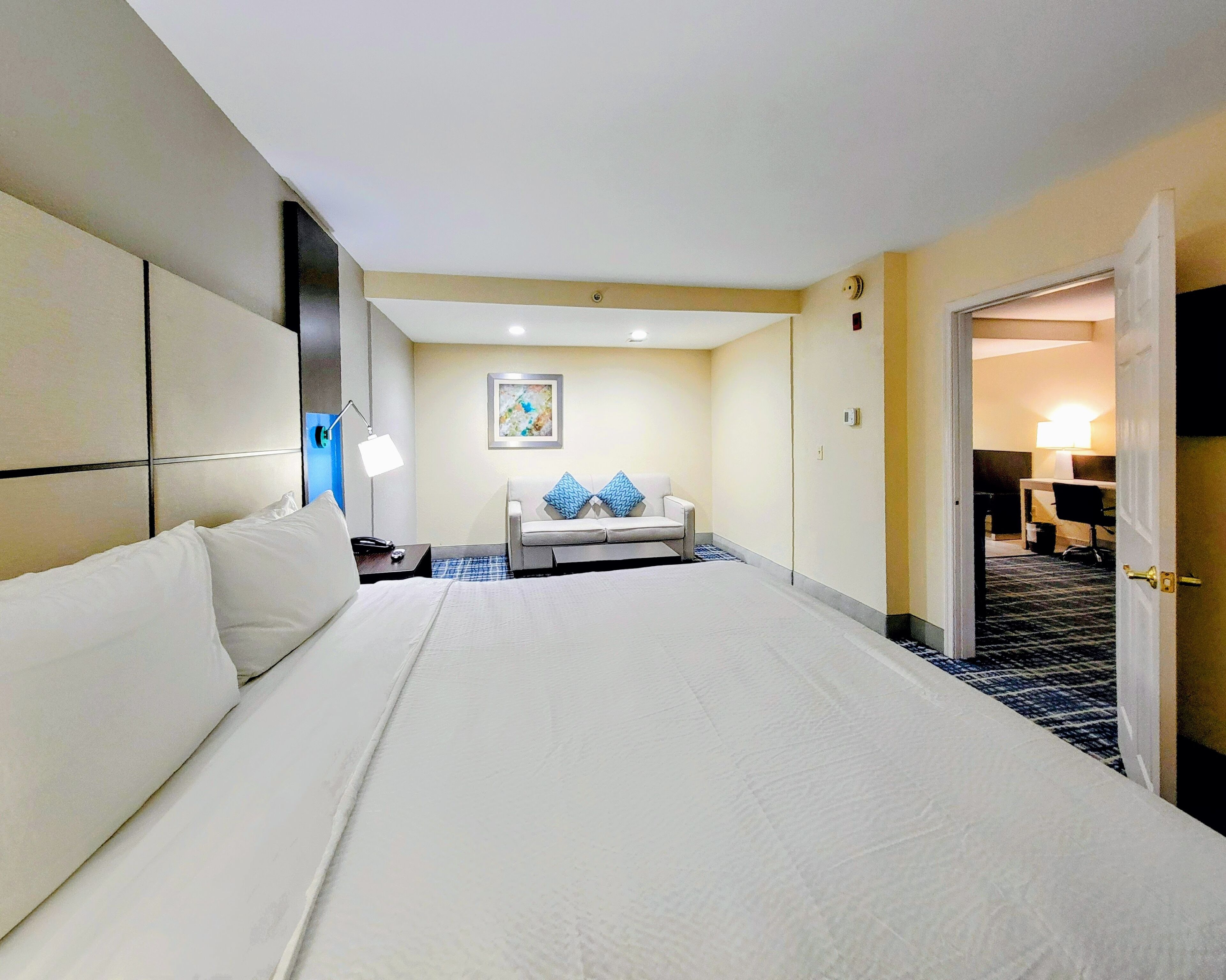 Una camera d'albergo con un letto grande, un comodino con una lampada, un divano con cuscini blu e un quadro appeso al muro.