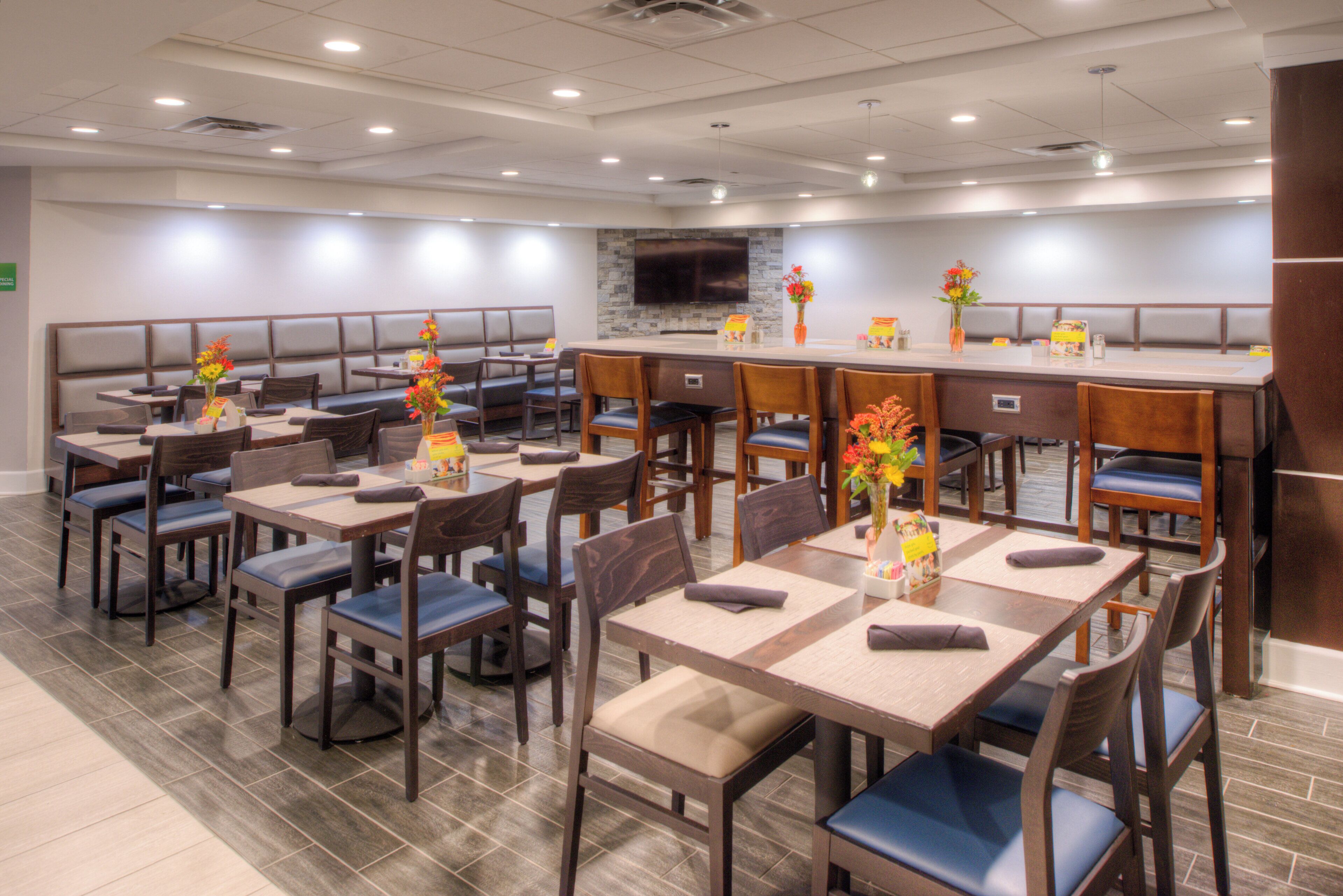 A hotel dining area with multiple tables and booths, a flat-screen TV mounted on the wall, and a variety of flowers in vases.