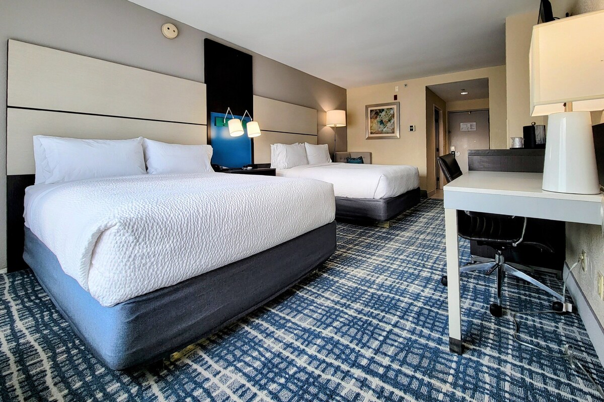 A hotel room with two beds, a desk with a lamp, a blue patterned carpet, and a painting on the wall.