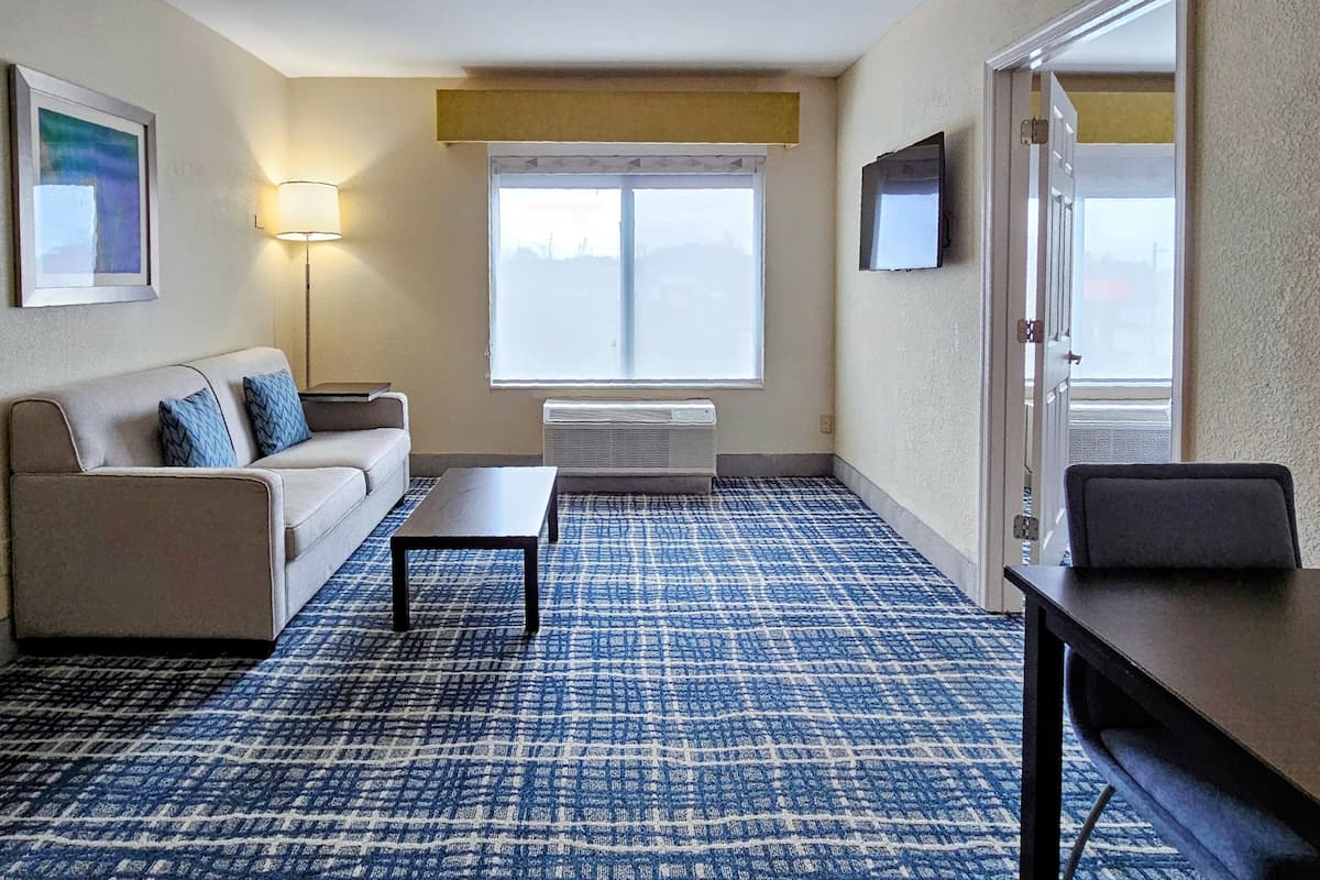 A hotel room with a sofa, a small table, a floor lamp, a television, and a large window.