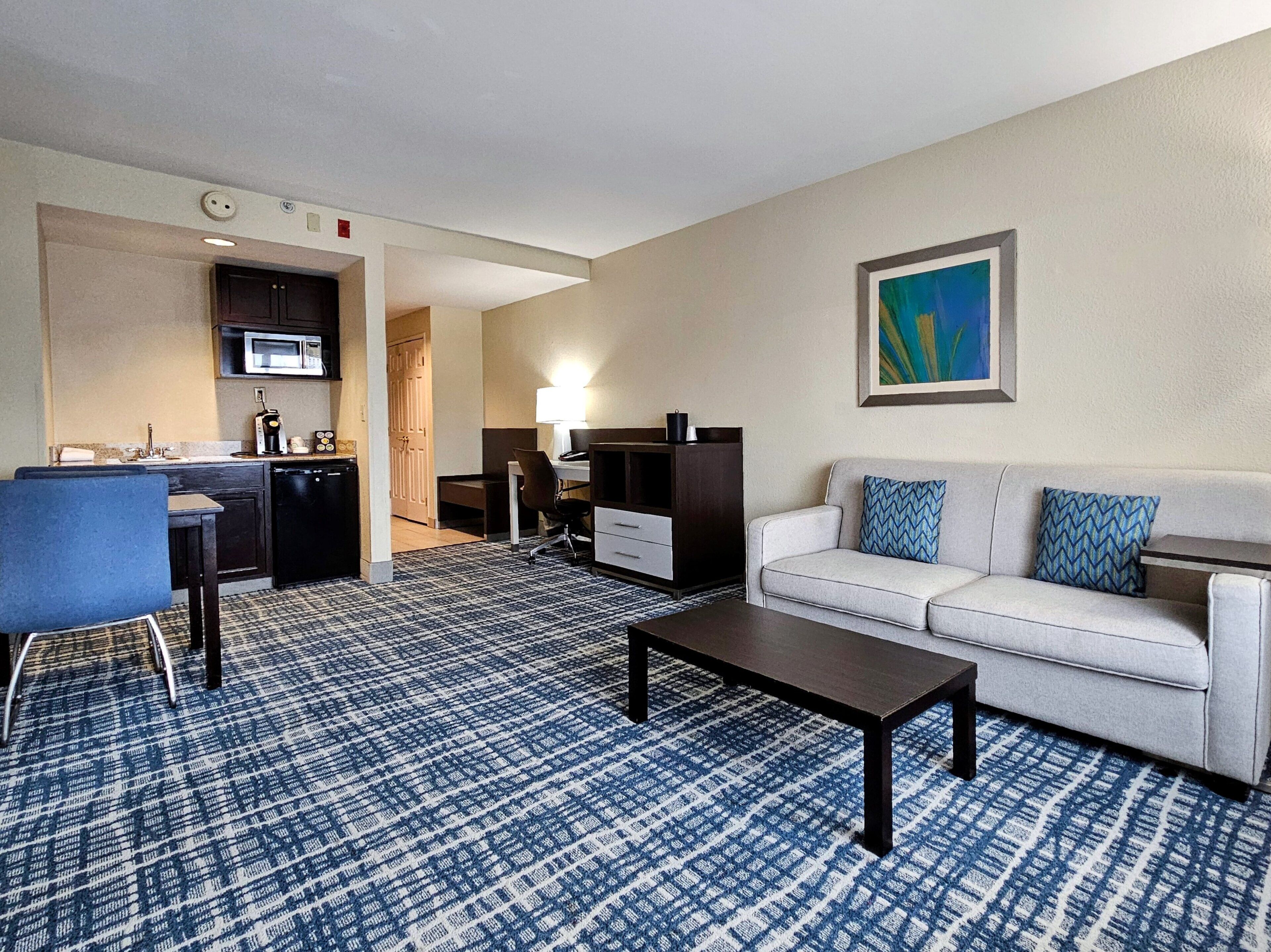 Una habitación de hotel con kitchenette, un comedor con mesa y sillas, un sofá con cojines estampados y un cuadro en la pared.