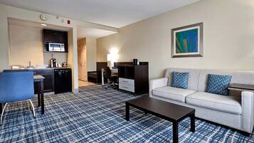 Una habitación de hotel con kitchenette, un comedor con mesa y sillas, un sofá con cojines estampados y un cuadro en la pared.