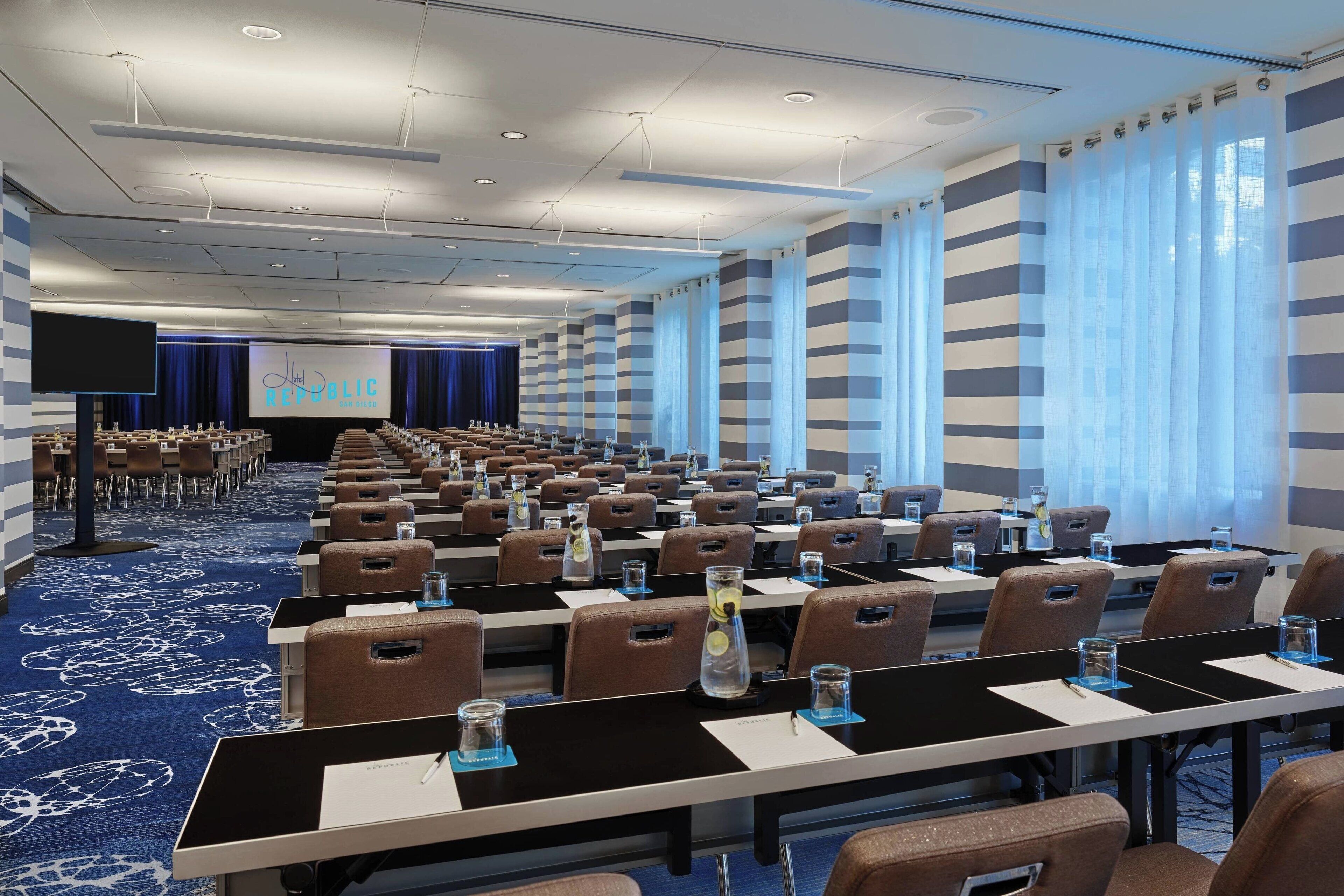 A conference room with rows of tables and chairs, a large screen at the front, and a blue carpet with a circular pattern.