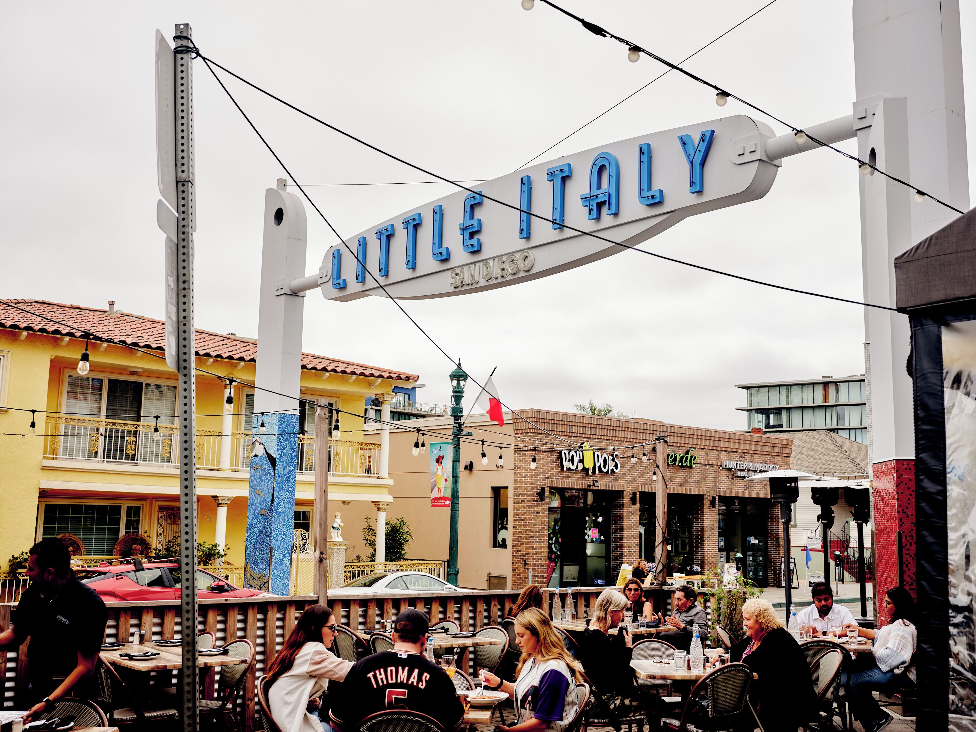 An outdoor dining area with tables and chairs, a sign reading "Little Italy," and various restaurant signs in the background.