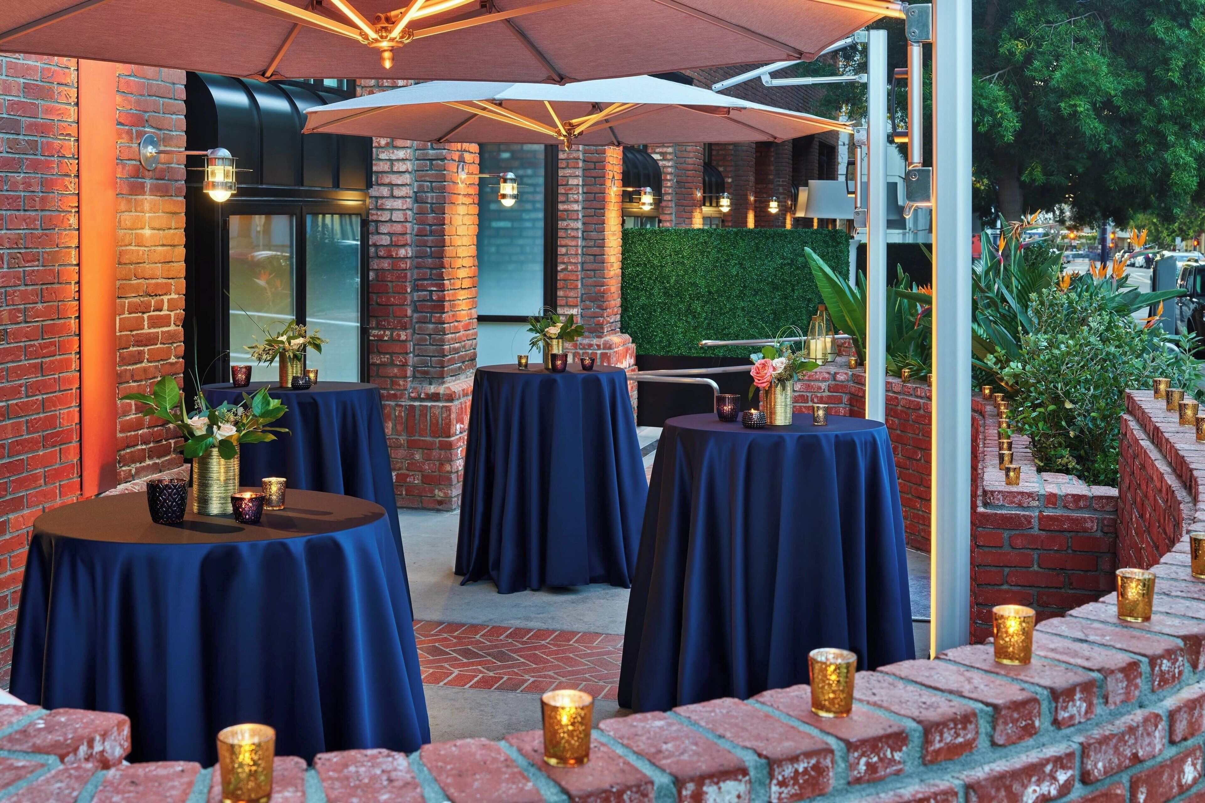 An outdoor seating area with round tables covered in blue tablecloths, surrounded by brick walls and greenery.