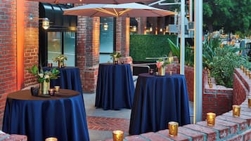 An outdoor seating area with round tables covered in blue tablecloths, surrounded by brick walls and greenery.