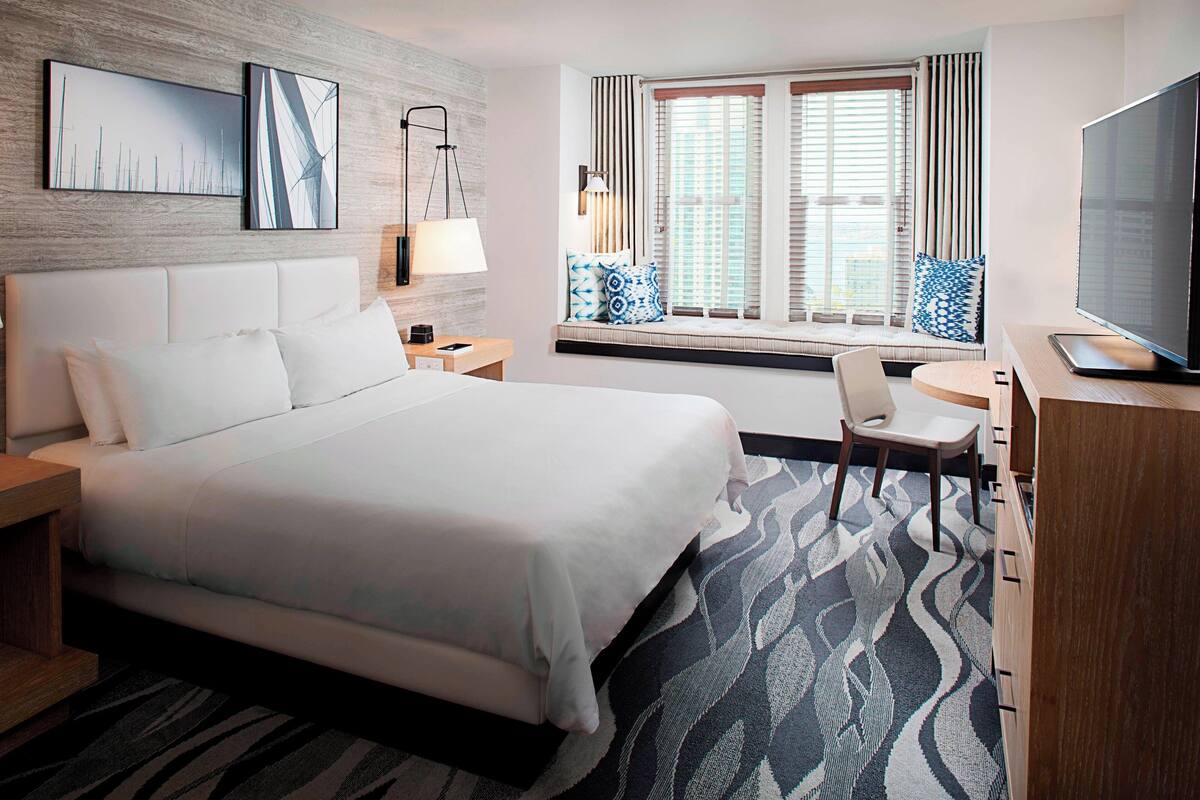 A modern hotel room with a large bed, a desk, a TV, and a window with blinds.