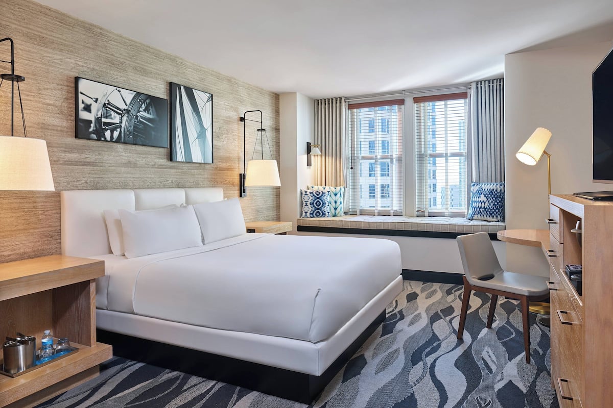 A modern hotel room with a large bed, a desk, and a chair. There are two windows with curtains, a wall-mounted TV, and artwork on the wall.