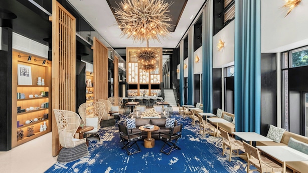 Un vestíbulo de hotel moderno con alfombra azul, separadores de madera y una zona de estar central con mesas y sillas.