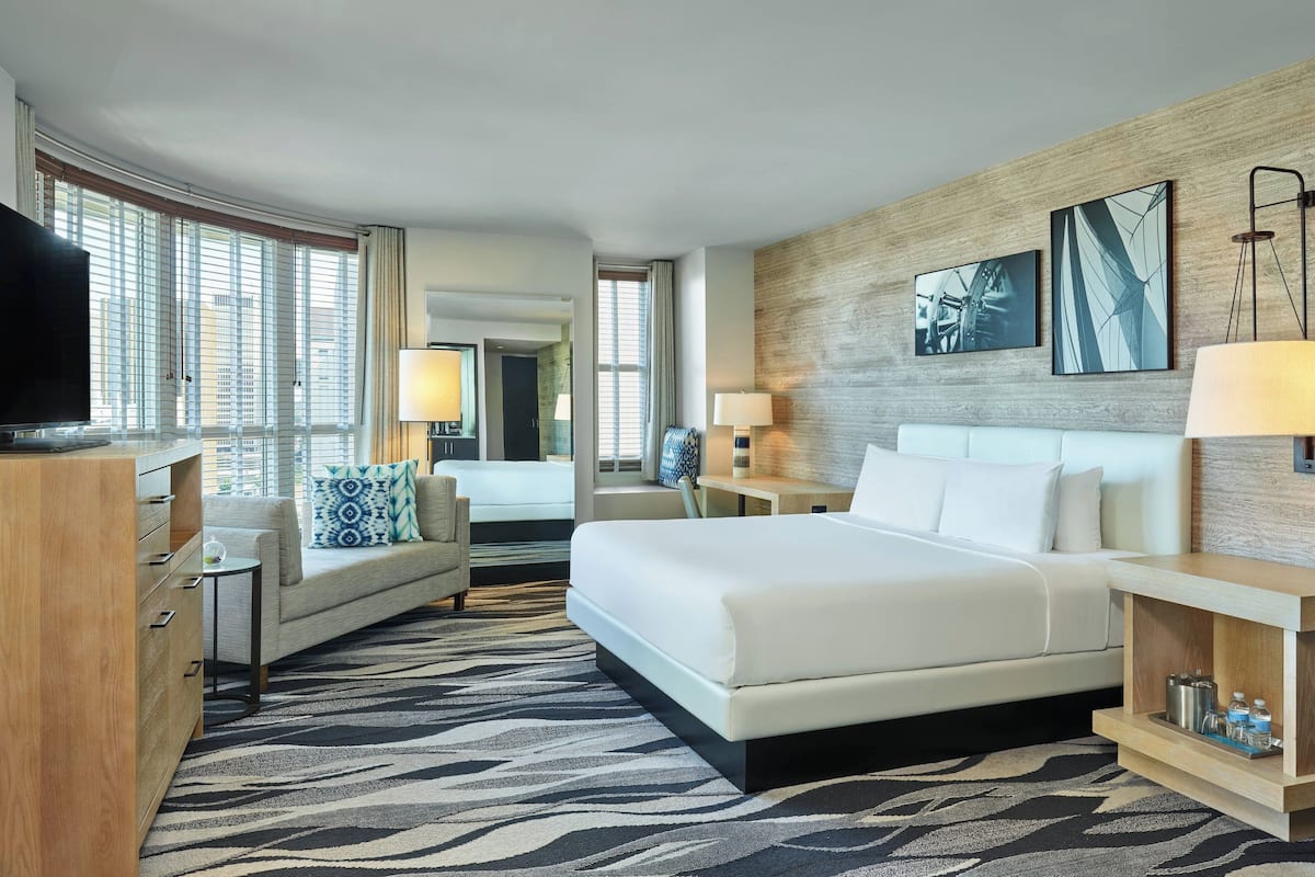 A hotel room with a large bed, a sofa, a TV, a desk, and a window with blinds.