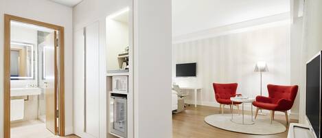 Una habitación de hotel moderna con suelo de madera, un tocador blanco con espejo, horno empotrado, televisor de pantalla plana, dos sillones rojos, una mesita redonda pequeña y un escritorio blanco con lámpara.