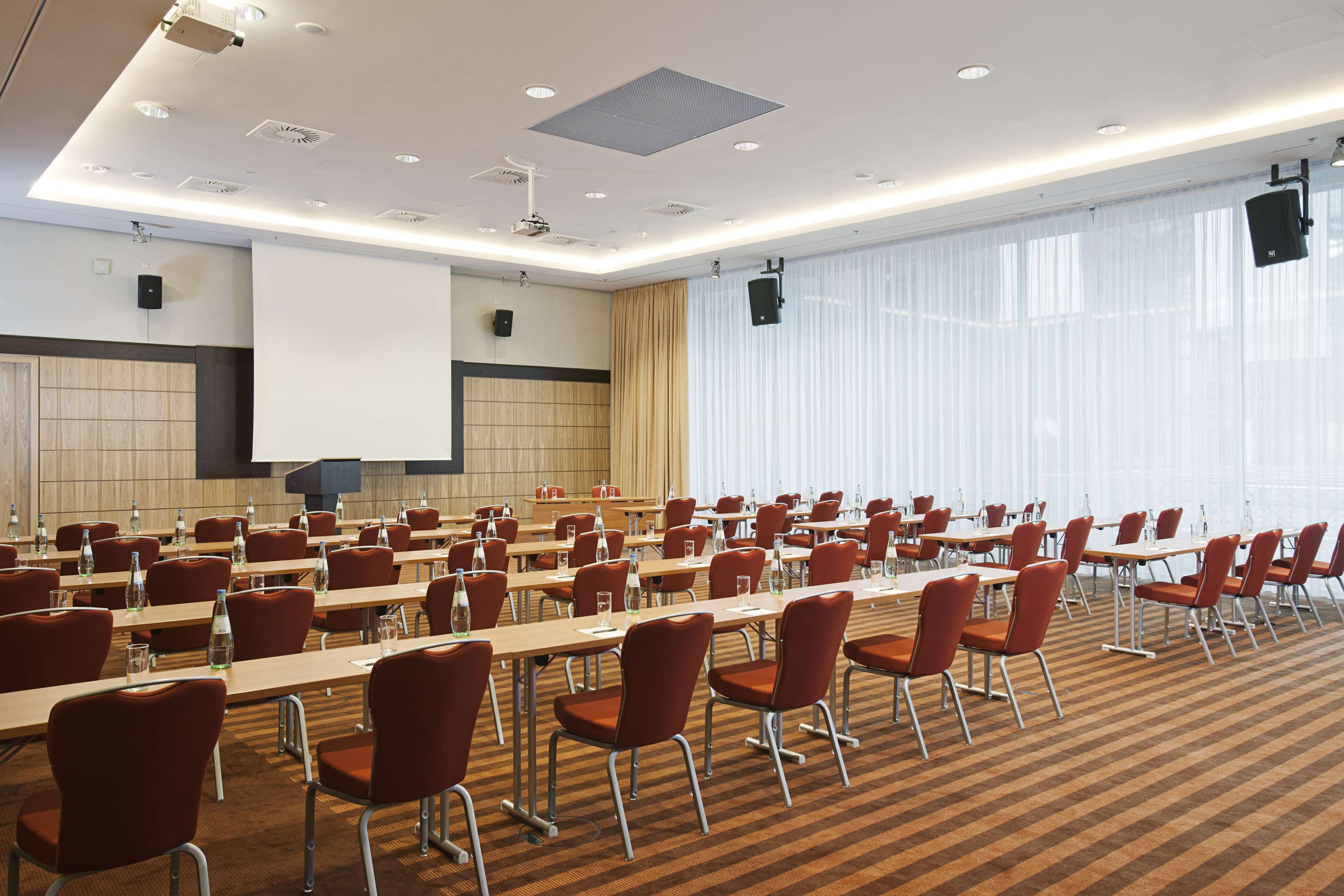 A conference room with rows of tables and chairs, a projector screen, and a large window with curtains.