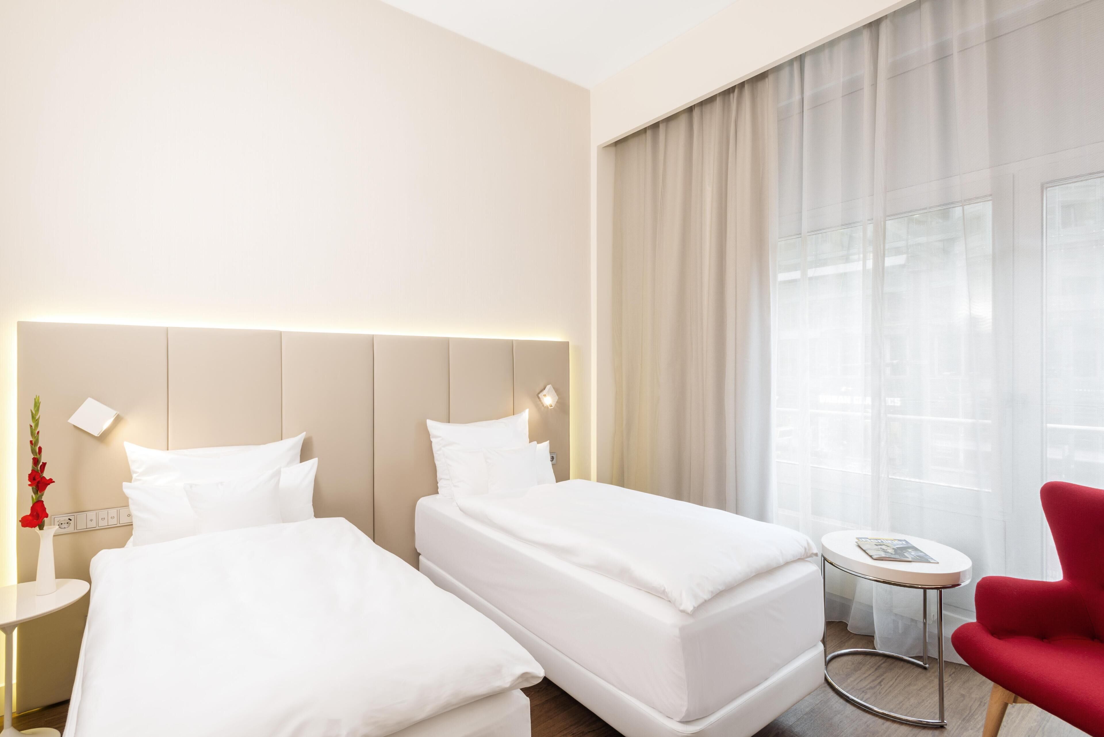 Hotellihuone, jossa on kaksi sänkyä, punainen tuoli, pieni pöytä ja ikkuna, jossa on verhot.