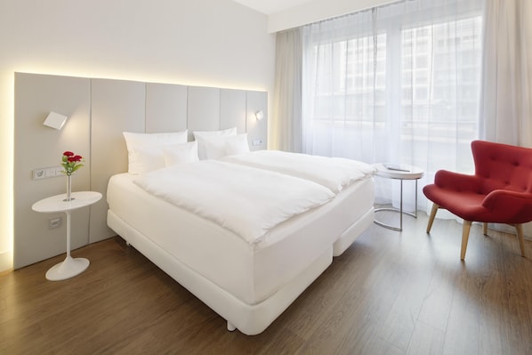A modern hotel room with a large bed, a small round table, a red chair, and a vase with flowers. - NH Collection Berlin Mitte Friedrichstrasse (Berlin)