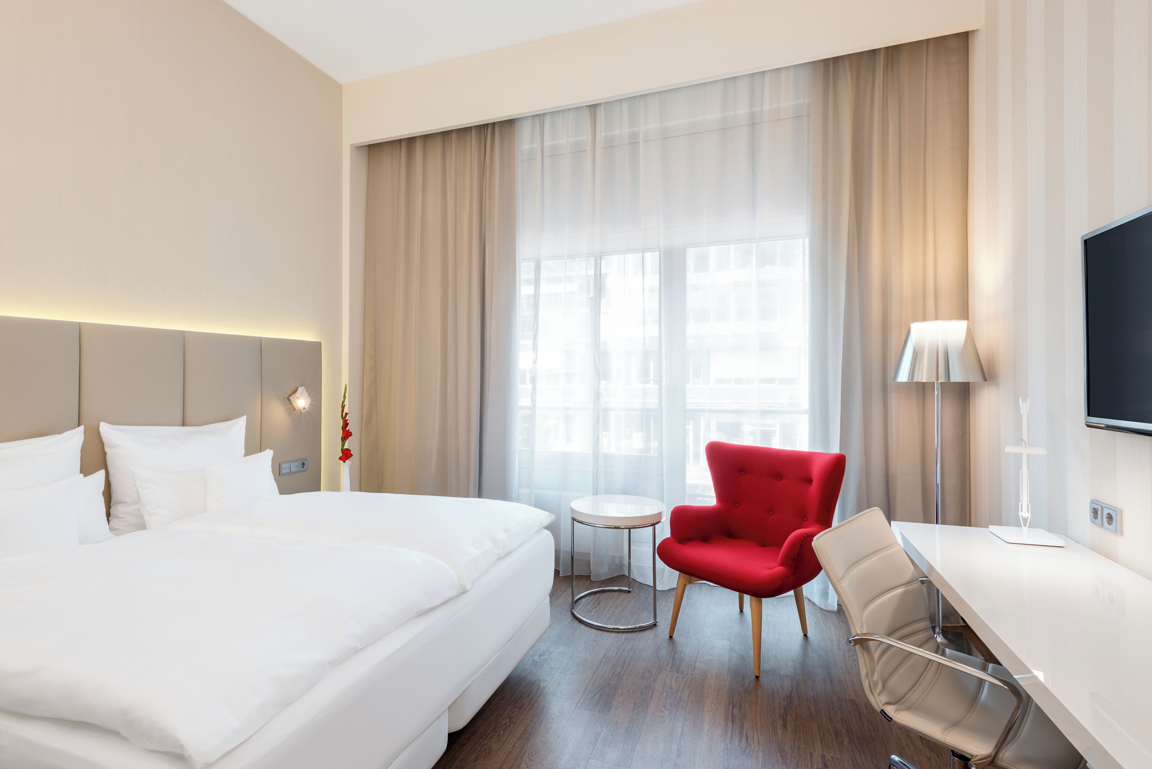 Habitación de hotel con una cama grande, una silla roja, una mesita redonda y un televisor montado en la pared.