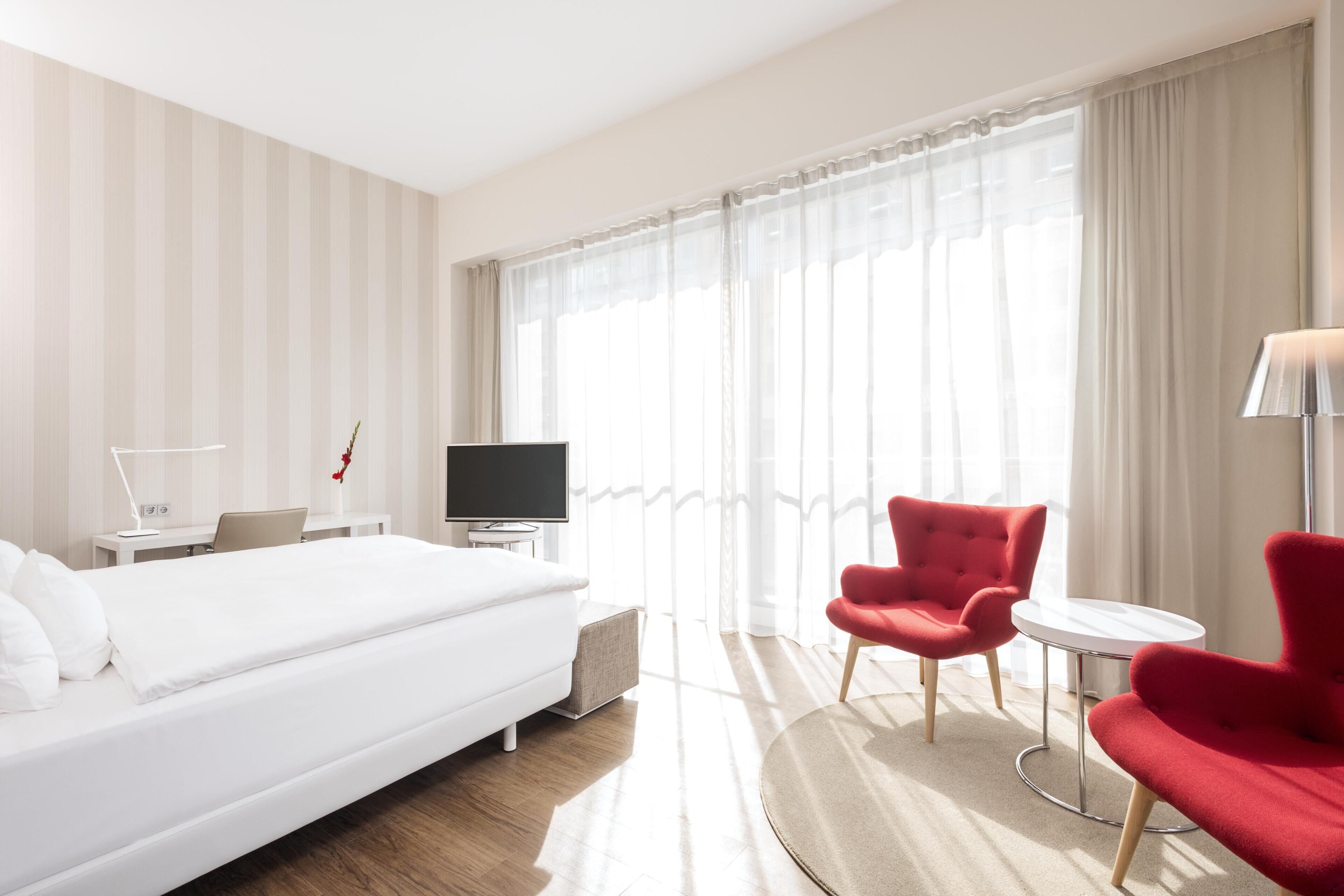 Habitación de hotel con una cama grande, un escritorio con una computadora portátil, un televisor, un sillón rojo y una mesita.