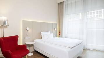 Habitación de hotel con una cama grande, un sillón rojo, una mesita redonda y un ventanal con cortinas traslúcidas.