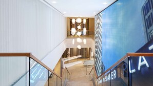 Un moderno vestíbulo de hotel con una escalera, una mesa de recepción y una gran pantalla digital que muestra la palabra "PLAT".