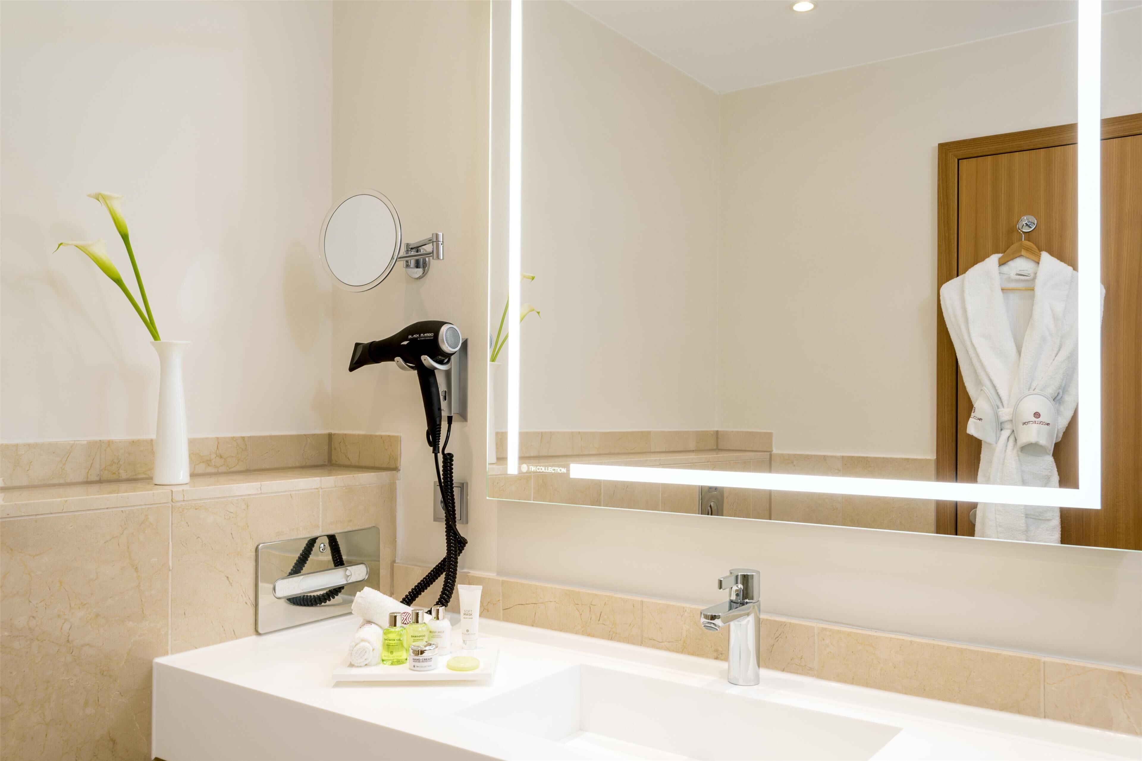 A modern bathroom with a white sink, a large mirror, a hairdryer, and a robe hanging on the door.