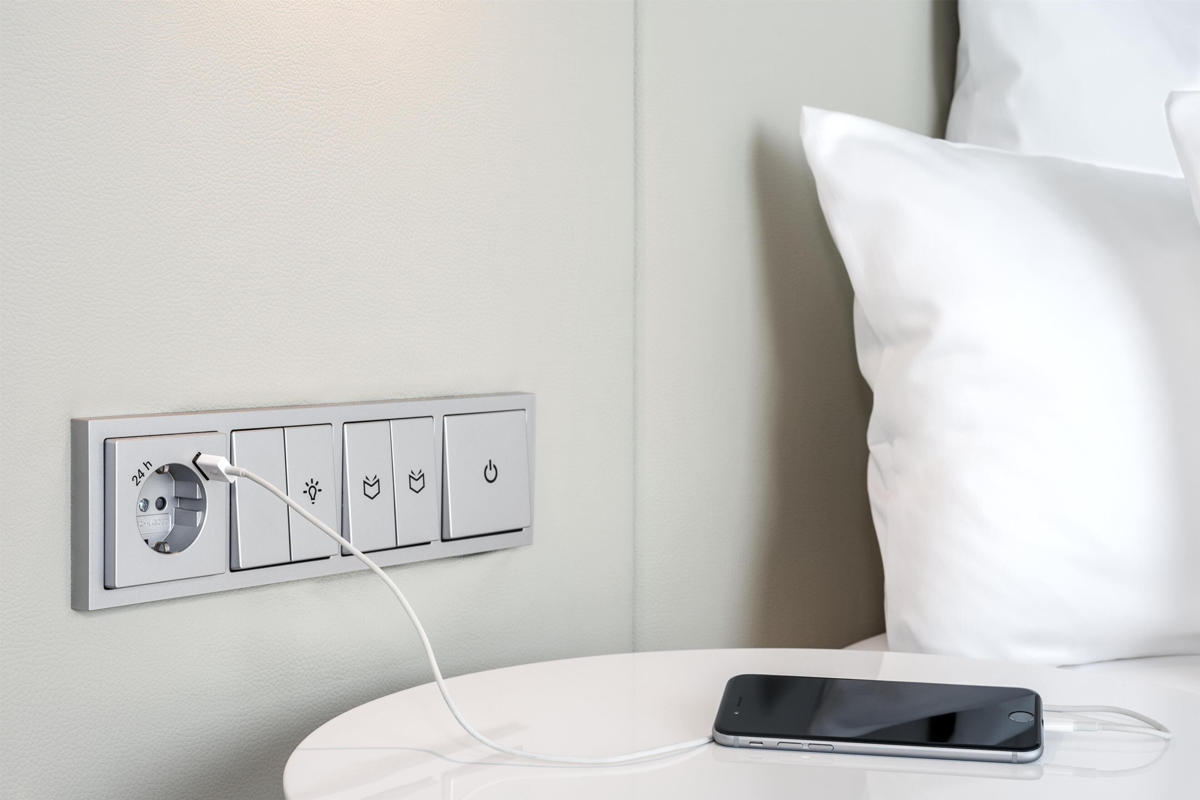 A white bedside table with a smartphone and charging cable, a wall socket with a power button, and a white pillow.