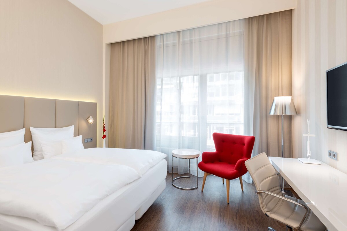 A hotel room with a large bed, a desk, a red chair, and a TV.