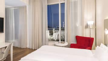 A hotel room with a large bed, a red armchair, a small round table, and a view of a tower through the window.