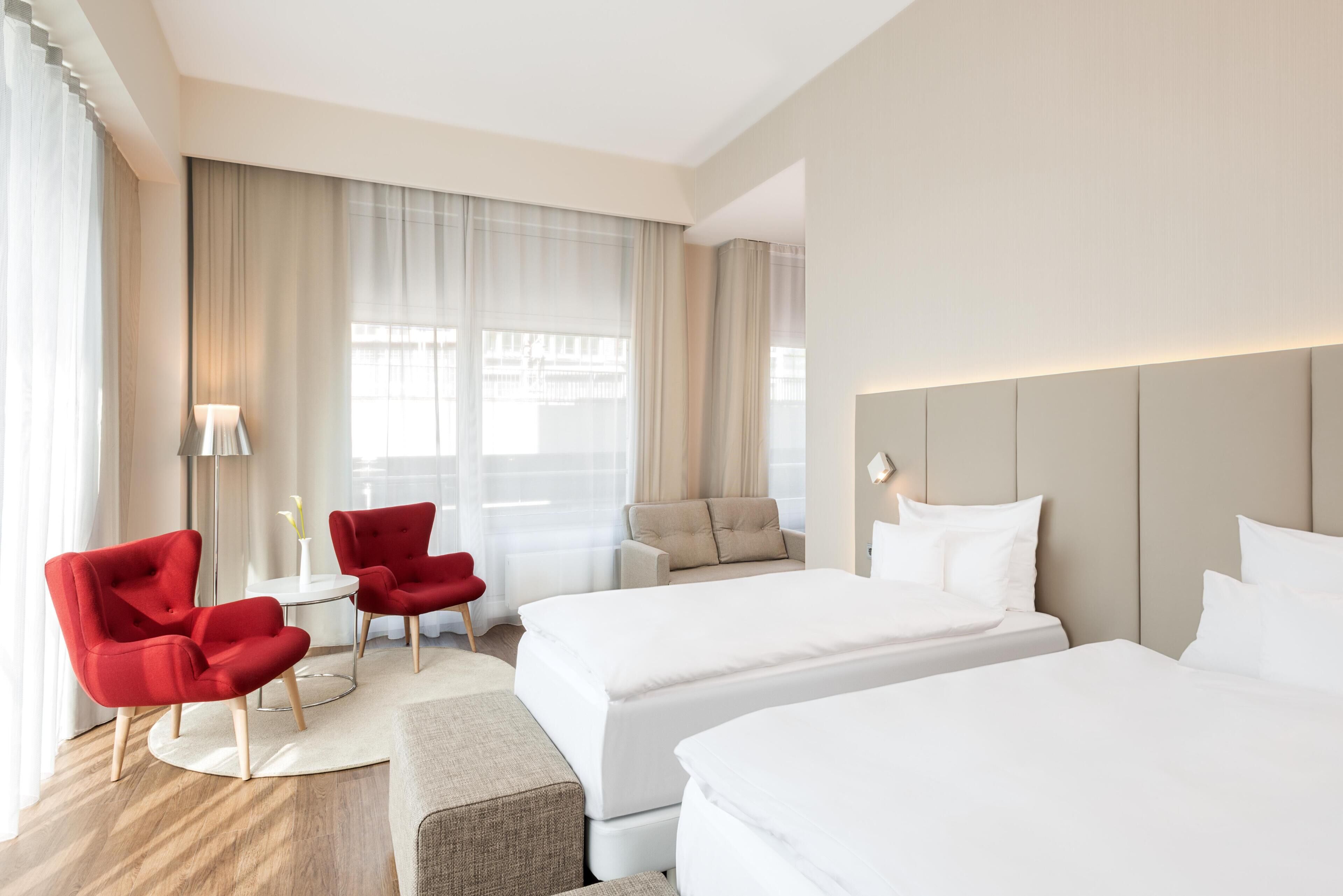 Una habitación de hotel con dos camas, un sillón rojo, un sofá color beige y una mesita con una lámpara.