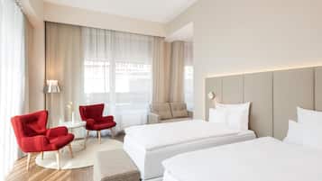 Una habitación de hotel con dos camas, un sillón rojo, un sofá color beige y una mesita con una lámpara.