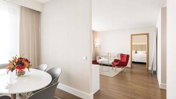 A modern hotel room with a dining area, a living space with a sofa and armchairs, and a large mirror.