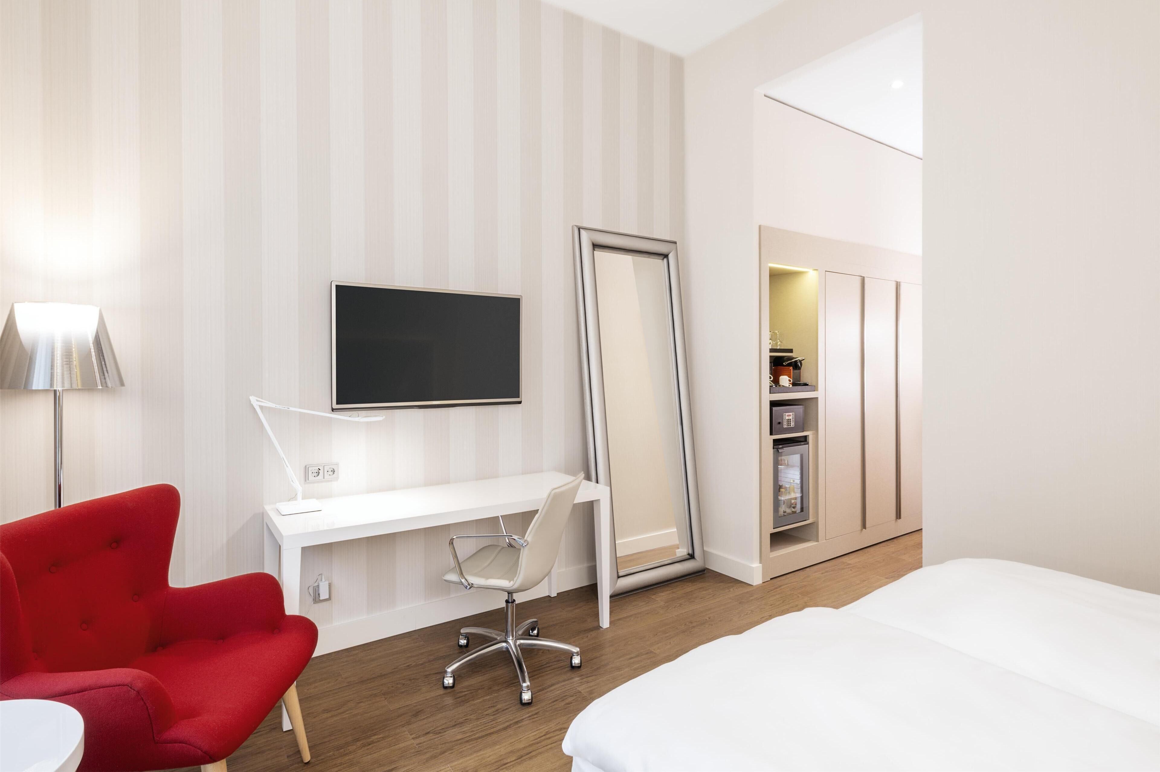 A hotel room with a white desk, a red chair, a bed, a TV mounted on the wall, and a wardrobe with built-in shelves.