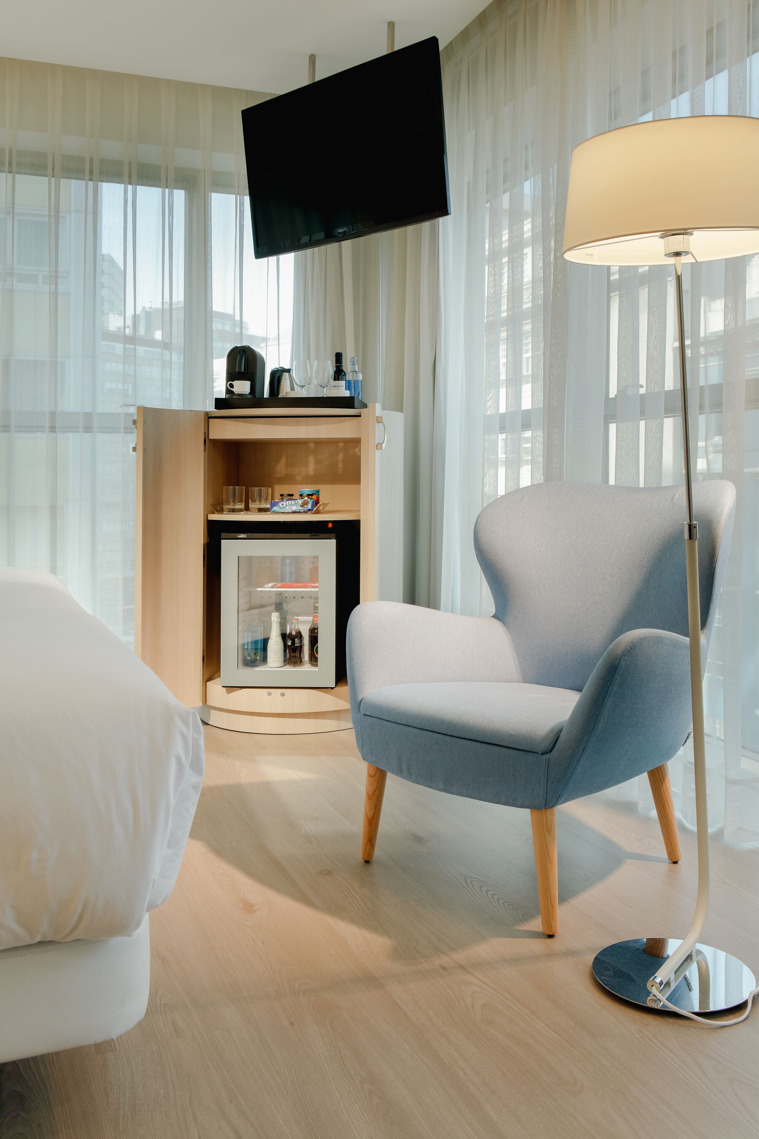 Una habitación de hotel moderna con un televisor de pantalla plana montado en la pared, una lámpara de pie, un minibar y un sillón cómodo.