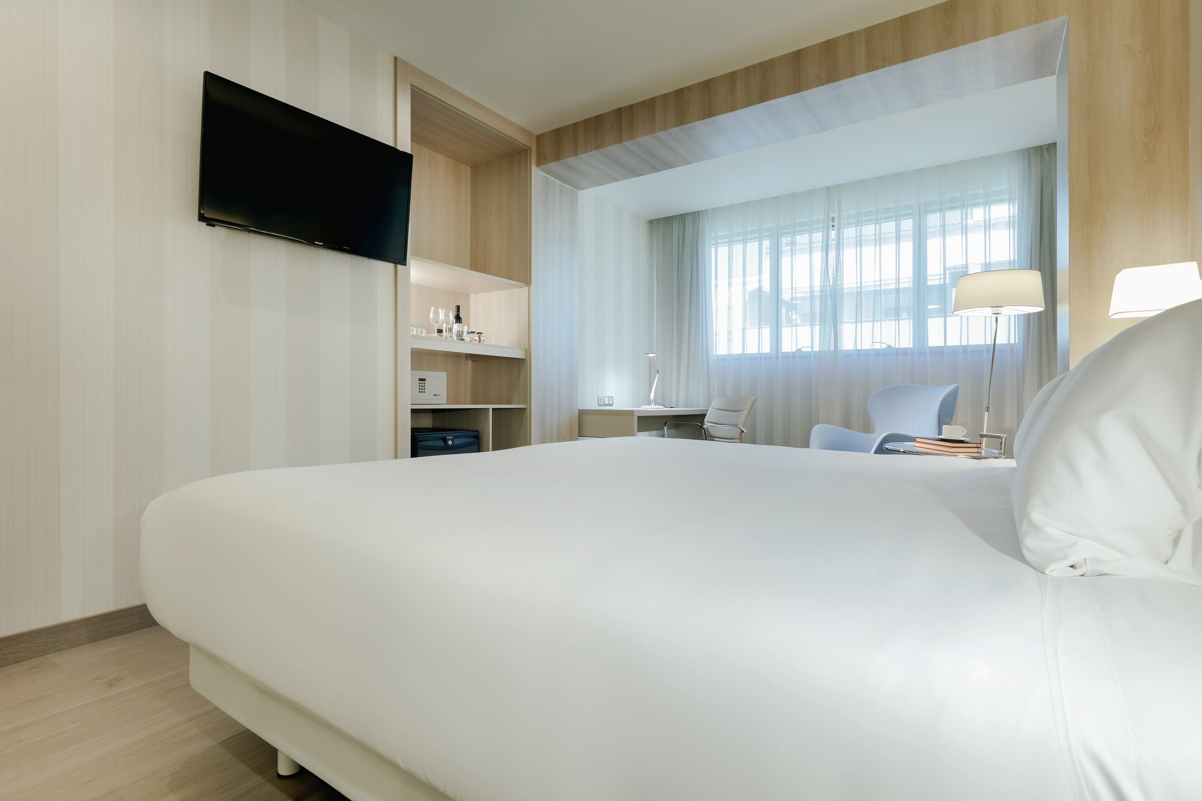 A modern hotel room with a large bed, a flat-screen TV mounted on the wall, a desk with a chair, and a window with sheer curtains.