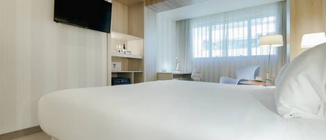 A modern hotel room with a large bed, a flat-screen TV mounted on the wall, a desk with a chair, and a window with sheer curtains.