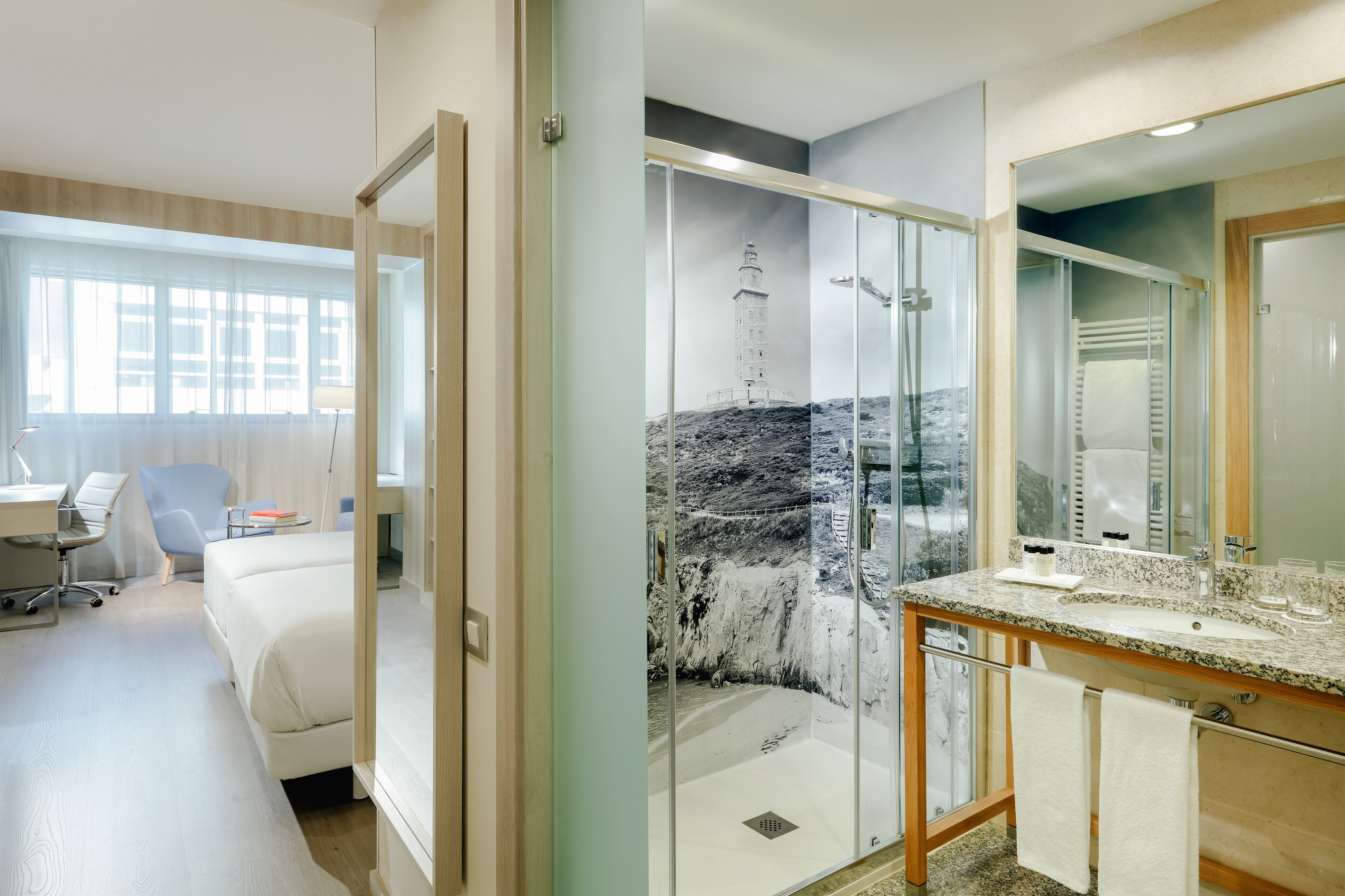 A hotel room with a bed, a desk, a chair, a bathroom with a sink and mirror, and a glass door leading to a scenic view of a lighthouse.