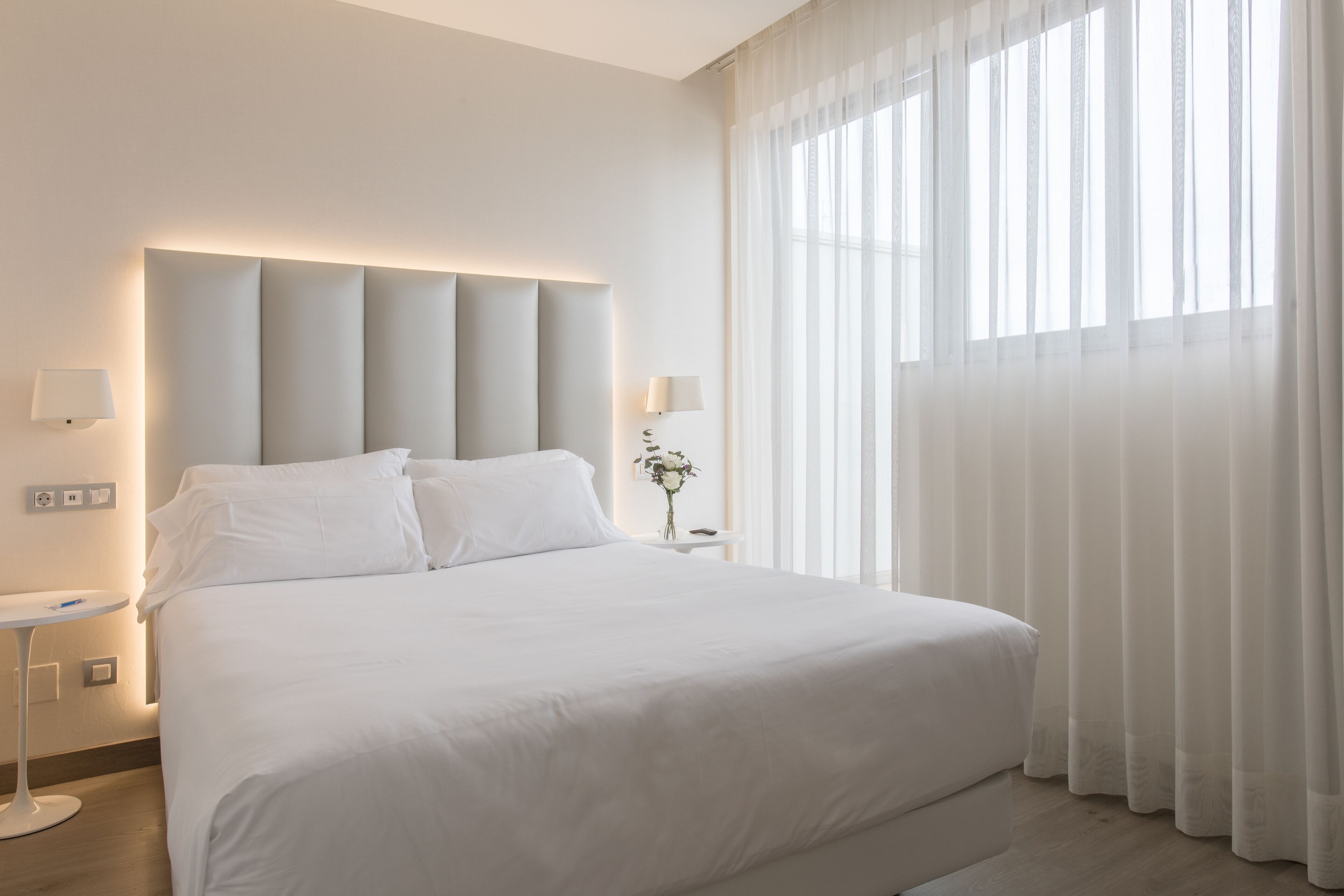 Una habitación de hotel moderna con una cama grande, dos mesitas de noche y un amplio ventanal con cortinas translúcidas.