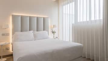 Una habitación de hotel moderna con una cama grande, dos mesitas de noche y un amplio ventanal con cortinas translúcidas.