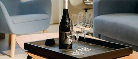 A modern hotel room with a wooden coffee table, a bottle of wine, and two glasses on a tray. There are two blue armchairs and a desk with a lamp in the background.