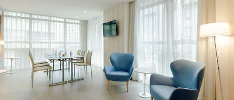 Quarto de hotel moderno com área de refeições, duas poltronas azuis, uma mesa pequena e uma TV fixada na parede.