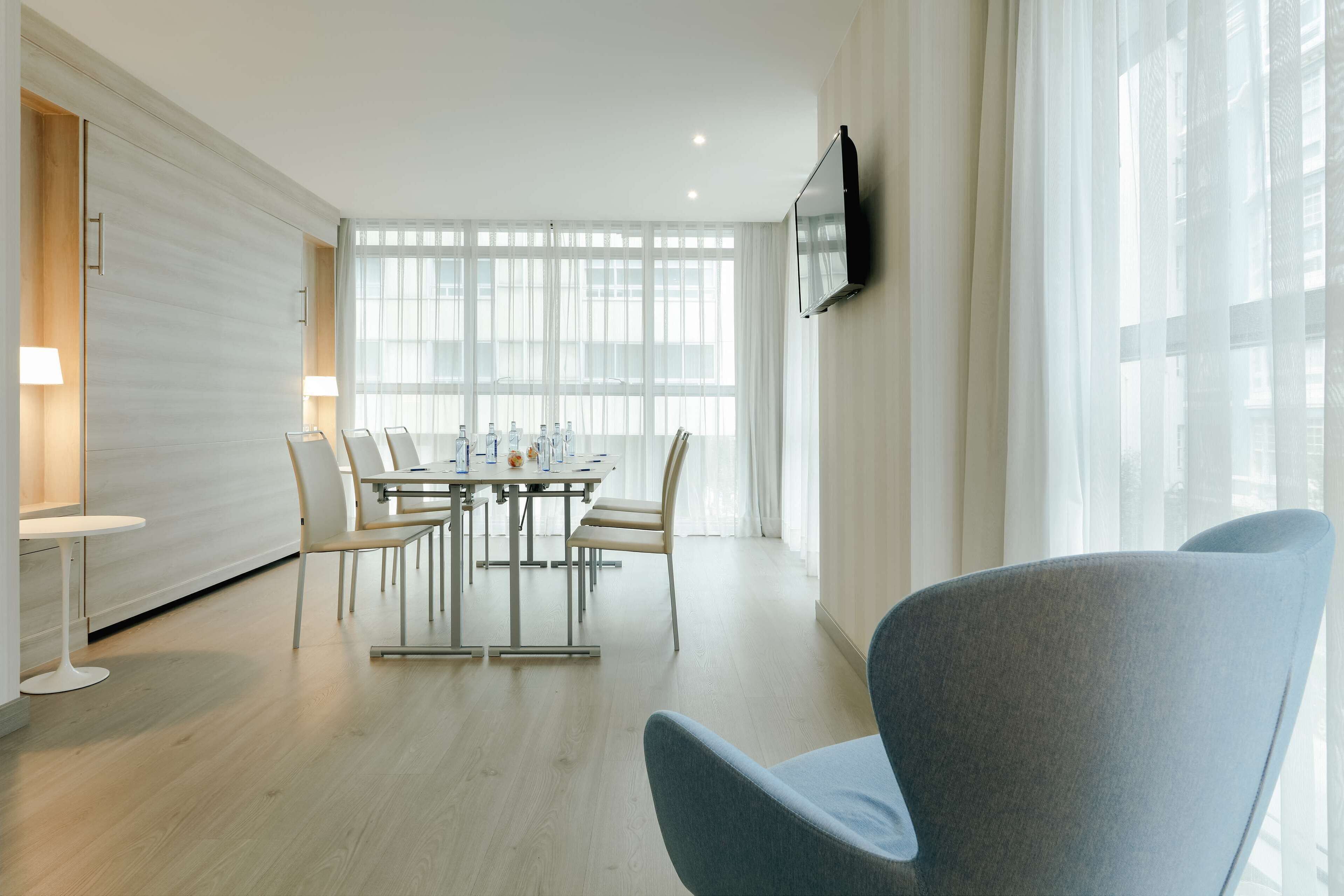 A modern hotel room with a dining area, large windows, and a flat-screen TV.