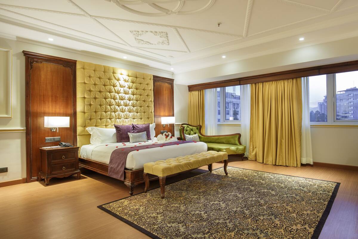 A spacious bedroom with a large bed, a wooden nightstand, a bench, and a view of the city through large windows.