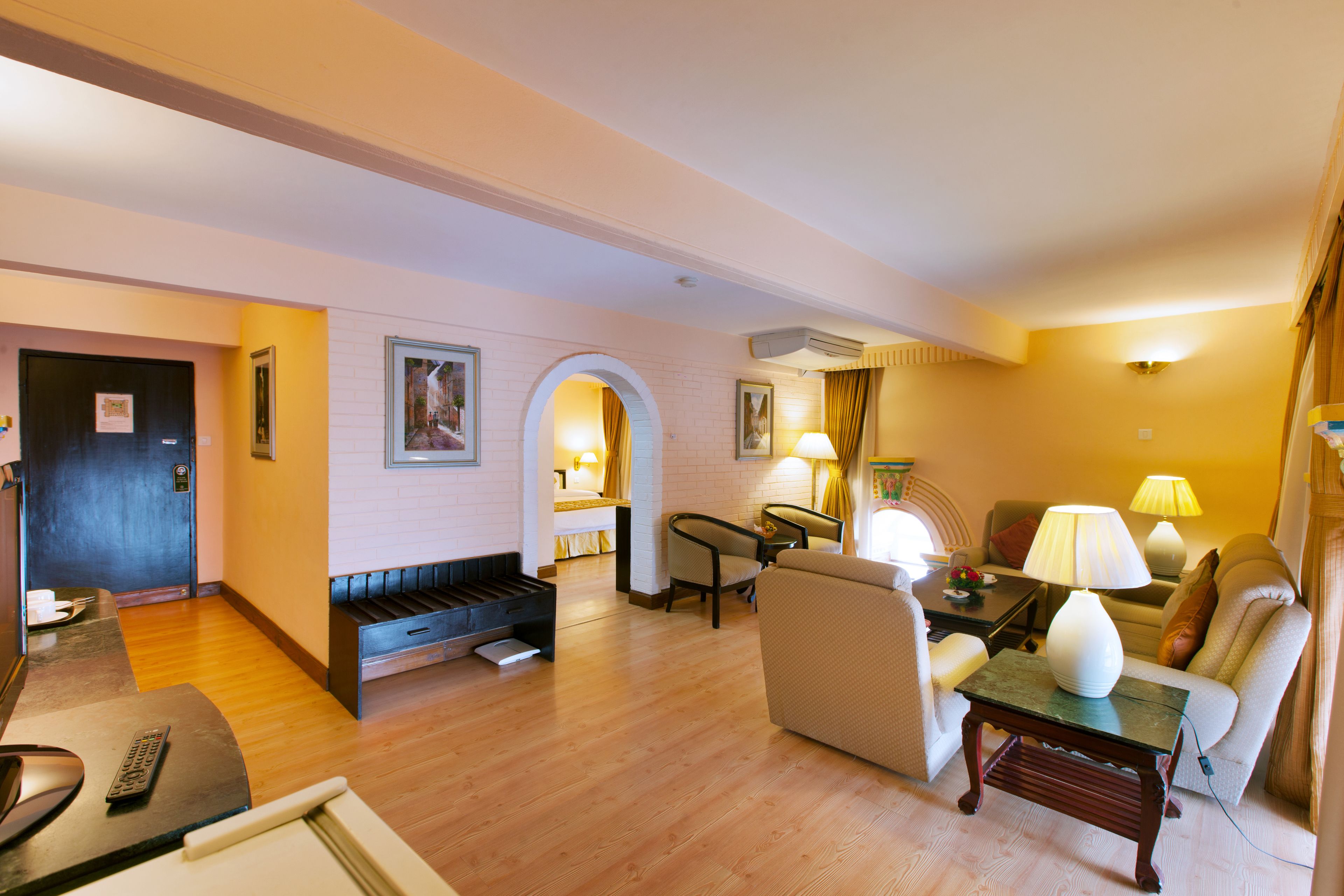 A hotel room with a wooden floor, a sofa, a chair, a small table with a lamp, and a bed with a headboard.