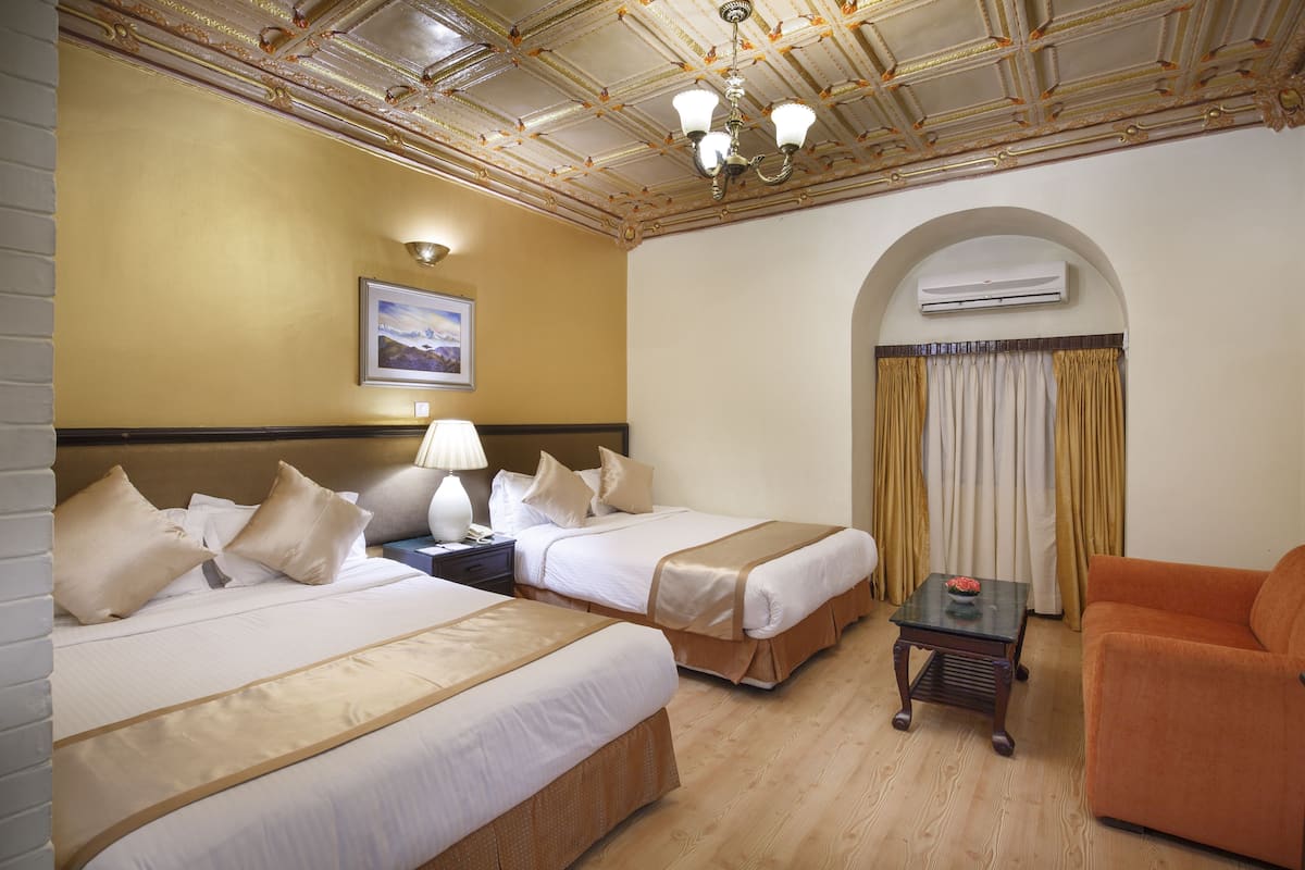 A hotel room with two beds, a sofa, a small table, an air conditioning unit, and a ceiling with ornate details.