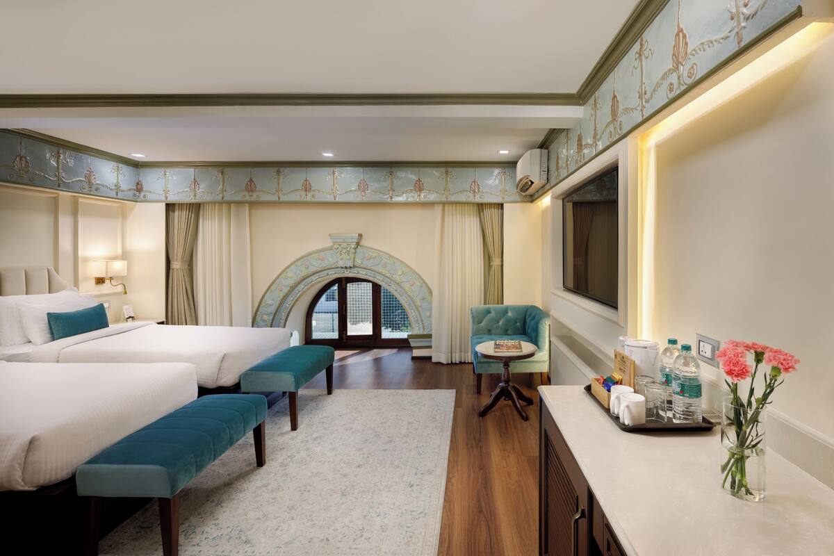 A hotel room with two beds, a sitting area with a table, a vase with flowers, and a large arched window.