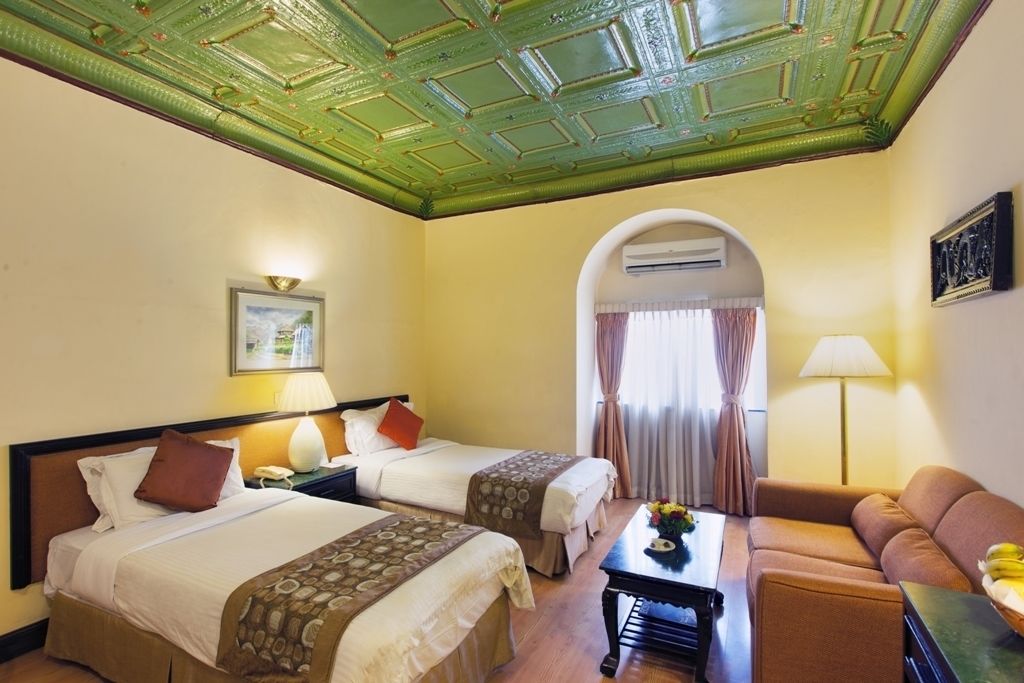 A hotel room with two beds, a sofa, a coffee table, and a ceiling with green decorative panels.
