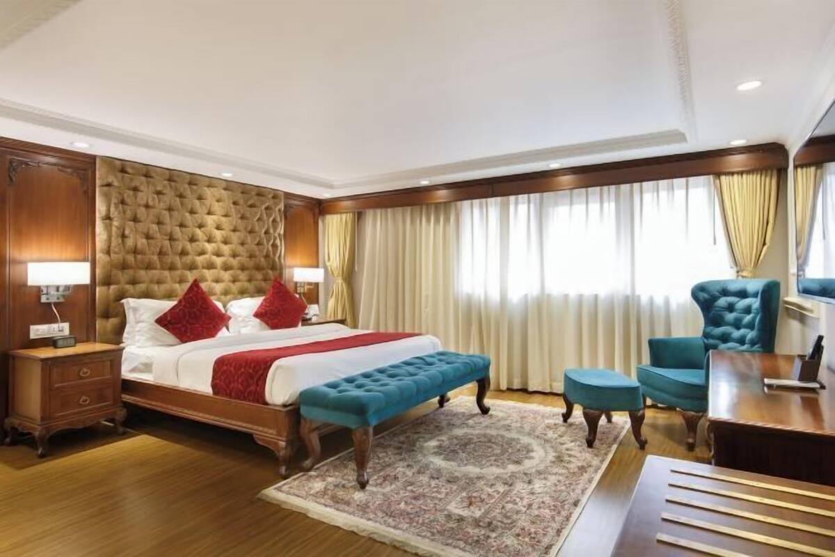 A hotel room with a large bed, a sitting area with a chair and ottoman, a desk, and a large window with curtains.