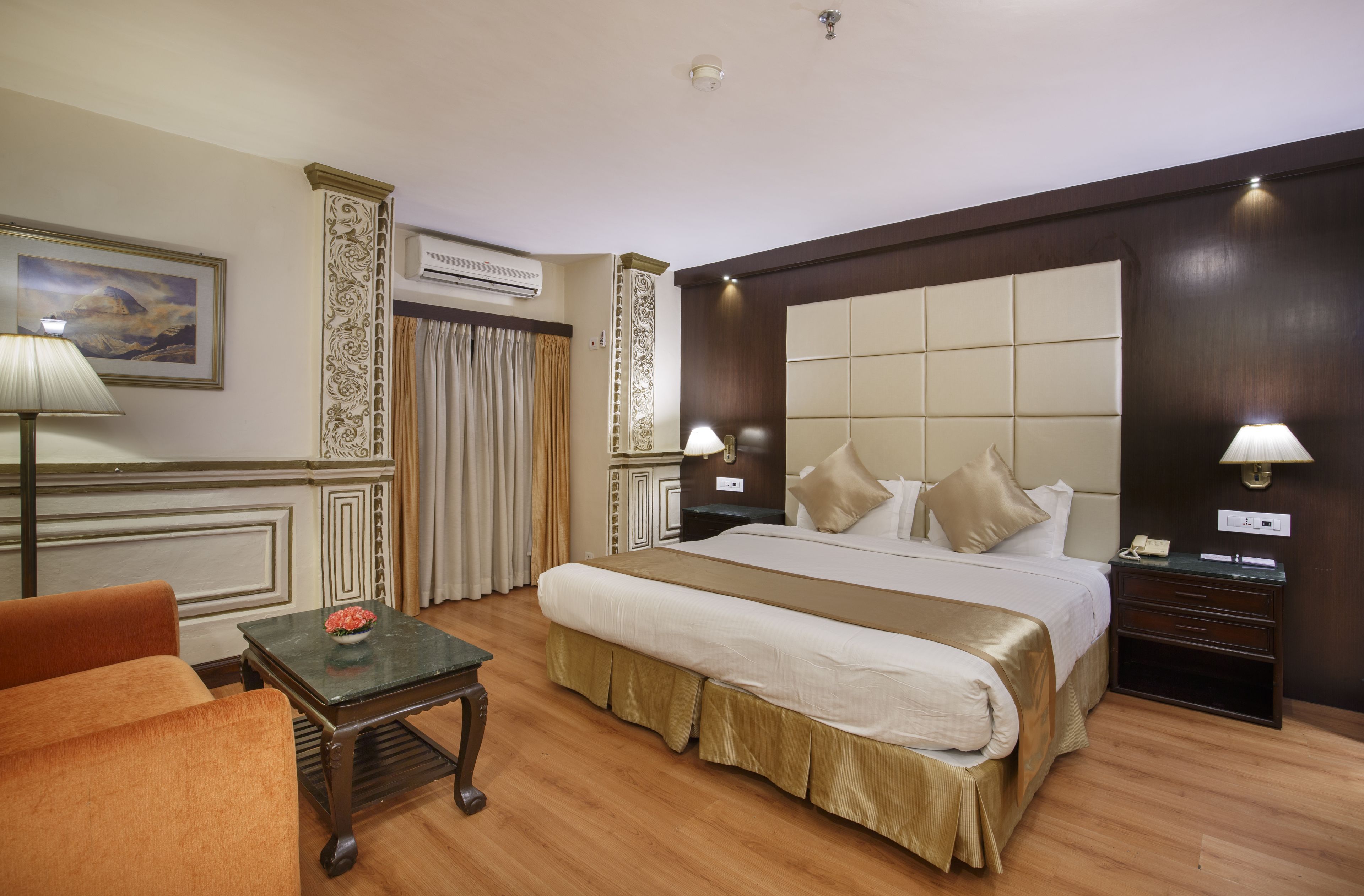 Una habitación de hotel con una cama grande, dos mesitas de noche con lámparas, una pequeña mesa con una flor, una silla y un equipo de aire acondicionado en la pared.